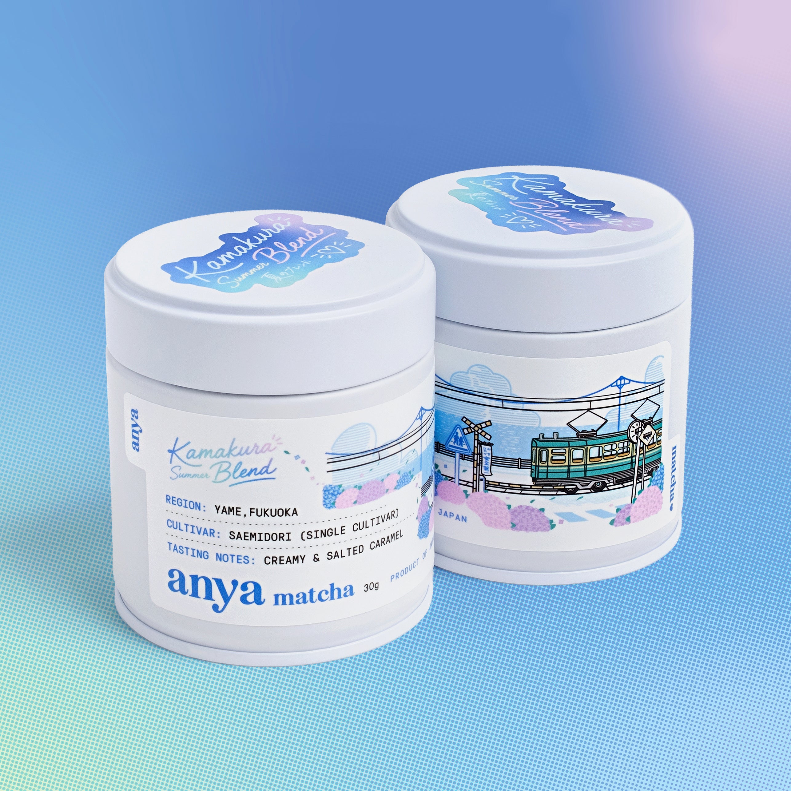 Two white cylindrical canisters of Anya Matcha Kamakura Blend matcha powder, decorated with blue and pink illustrations of a streetcar and cherry blossoms, set against a blue gradient background.